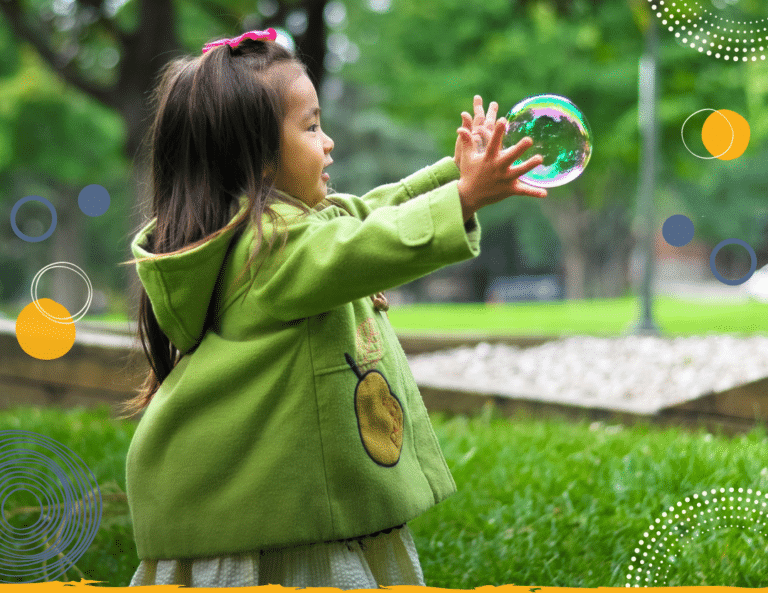 Photo of a young girl in a green coat with her arms outstretched trying to catch a bubble in a park to illustrate how play helps children's mental health