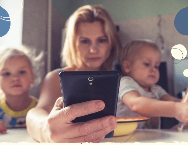 Photo of mum loking at her phone sitting at a table with two young children to illustrate article on how being a working parent impacts children according to research