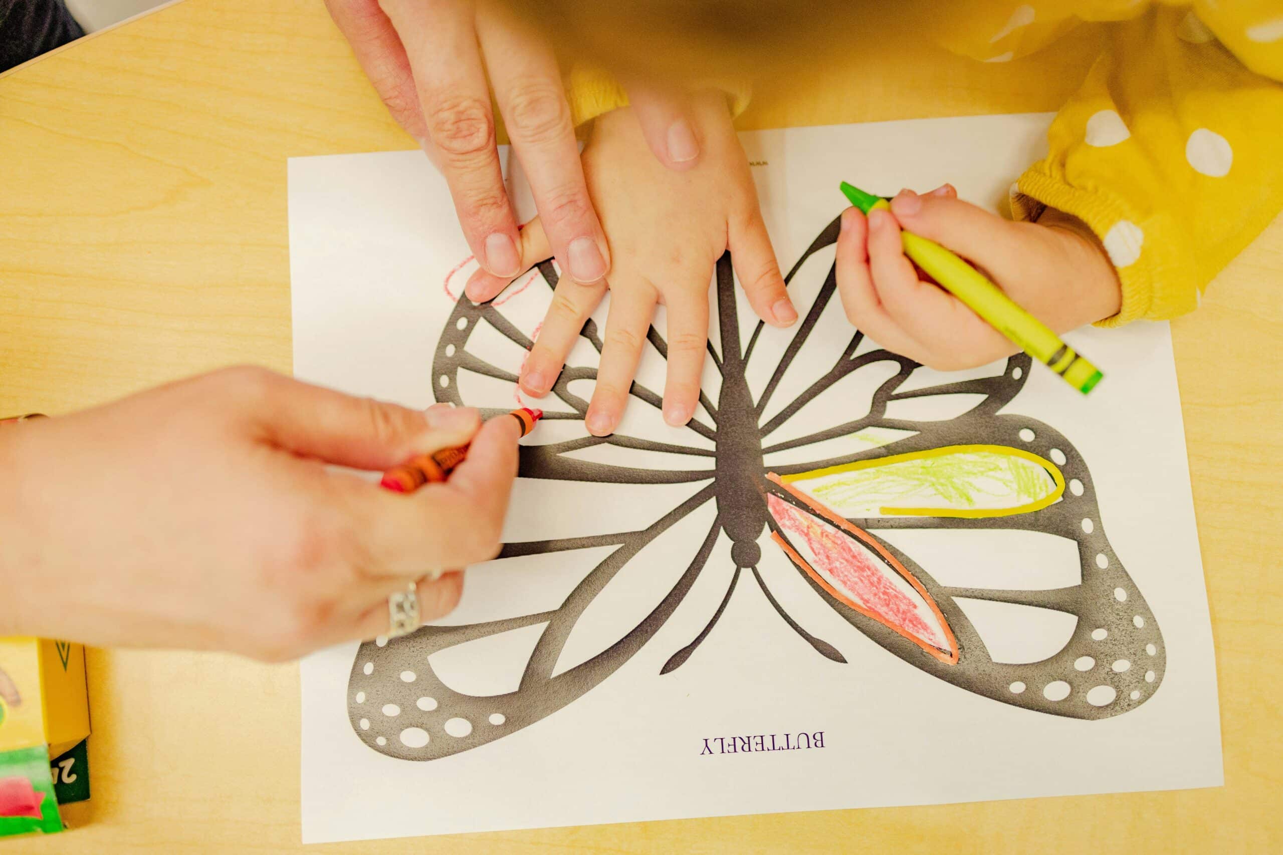 Photo of young child colouring in a butterfly drawing assisted by an adult