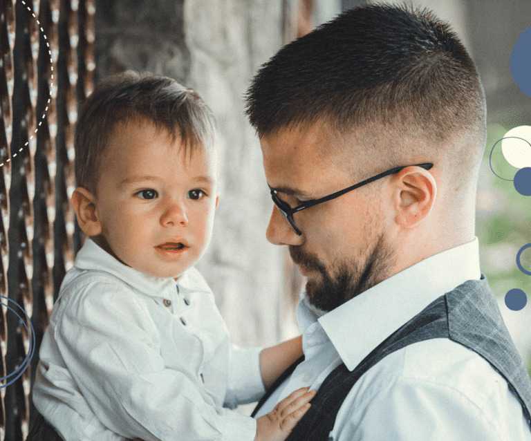 photo of white man with spectacles and a beard smartly dressed in a white shirt and grey waistcoat and holding a toddler, to illustrate article on how companies can support working parents